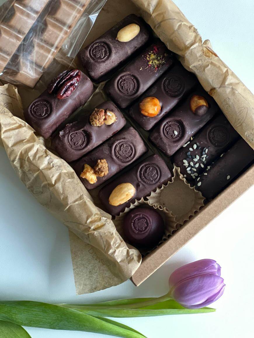 An open box of assorted dark chocolate candies topped with various nuts and seeds, sitting on crumpled brown paper next to a single purple tulip on a white surface.