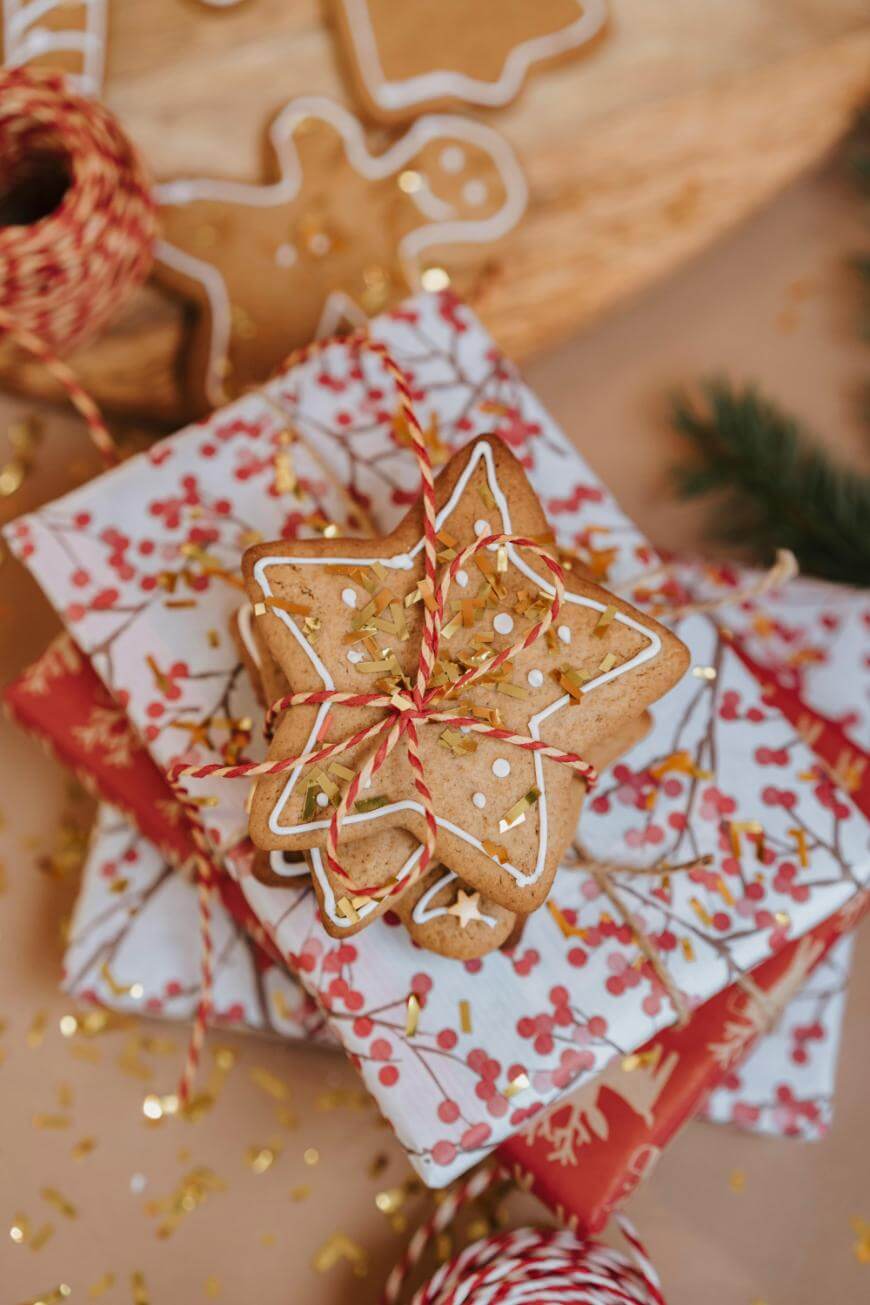 A stack of wrapped gifts decorated with festive paper and topped with star-shaped iced cookies, tied with red and white string, surrounded by gold confetti and a spool of string.