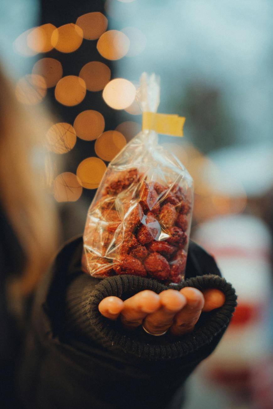 A person wearing a dark sleeve holds out a clear plastic bag filled with roasted nuts. Warm, blurred lights create a festive background, and the focus is on the hand and the bag of nuts.