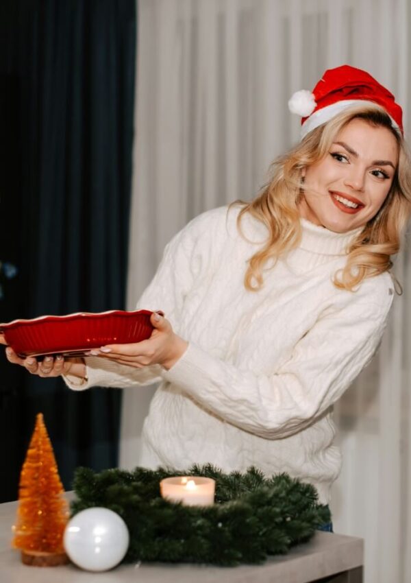 A smiling woman in a Santa hat and white sweater holds a red baking dish. A lit candle, a gold Christmas tree, garland, and a white ornament are on the table in front of her. She stands indoors with curtains behind her.