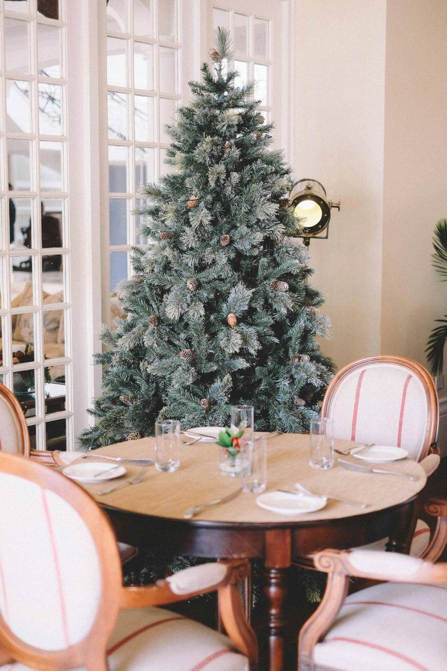 A decorated Christmas tree stands next to a round dining table set with plates, glasses, and napkins. Four upholstered chairs surround the table in a cozy, well-lit room with large windows in the background.