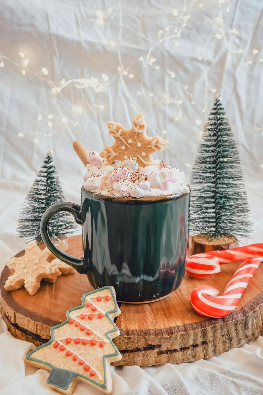 A mug of hot chocolate topped with marshmallows and a snowflake cookie sits on a wooden tray, surrounded by decorated cookies, candy canes, and small Christmas trees with twinkling lights in the background.