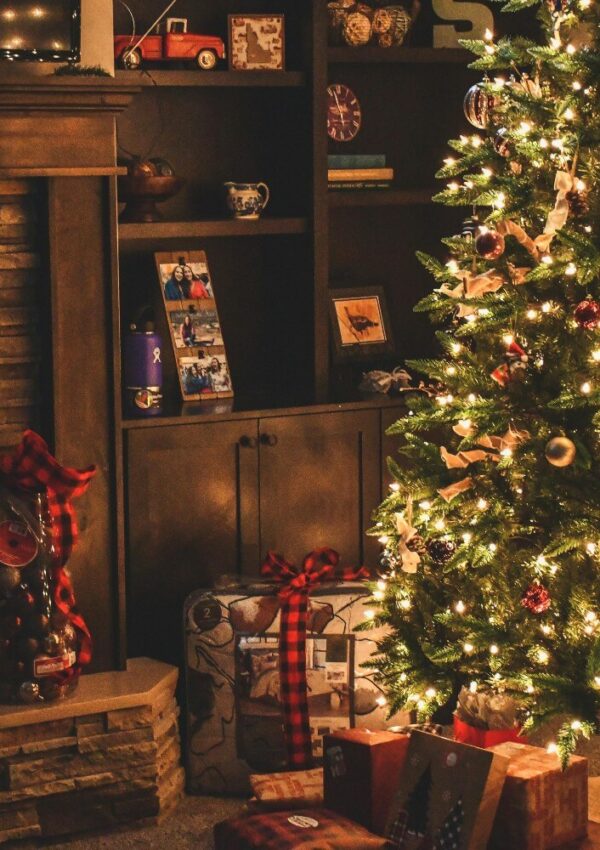 A warmly lit living room decorated for Christmas, featuring a glowing Christmas tree, wrapped presents underneath, a fireplace, and shelves with framed photos, ornaments, and holiday decor.