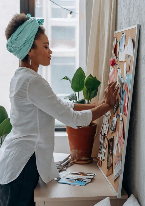 A woman wearing a white shirt and green headscarf arranges photos and papers on a bulletin board next to a window, with a potted plant nearby.