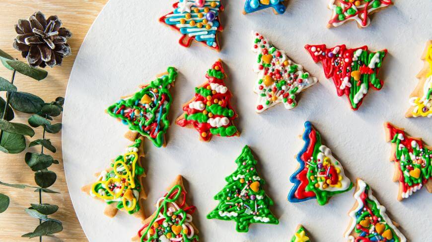 Colorfully decorated Christmas tree-shaped cookies are arranged on a white plate. The cookies are adorned with icing and sprinkles. Eucalyptus leaves and a pinecone are placed nearby for decoration.