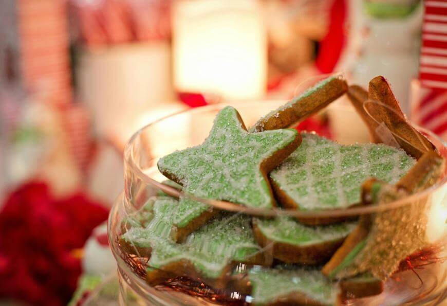 A glass bowl filled with star-shaped cookies decorated with green icing and sugar, set on a festive table with red, white, and soft lights in the background.