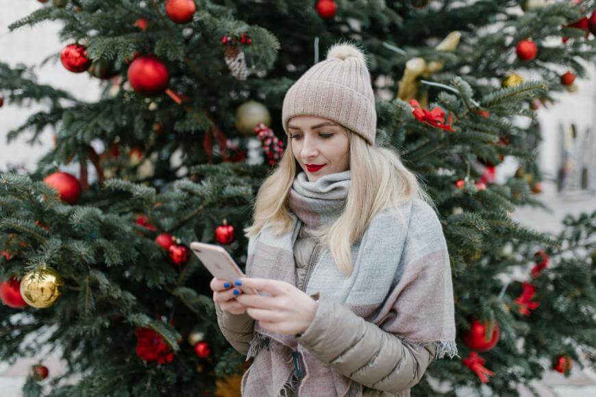 A woman in a beige hat and scarf stands in front of a decorated Christmas tree, looking down at her smartphone. The tree is adorned with red and gold ornaments.