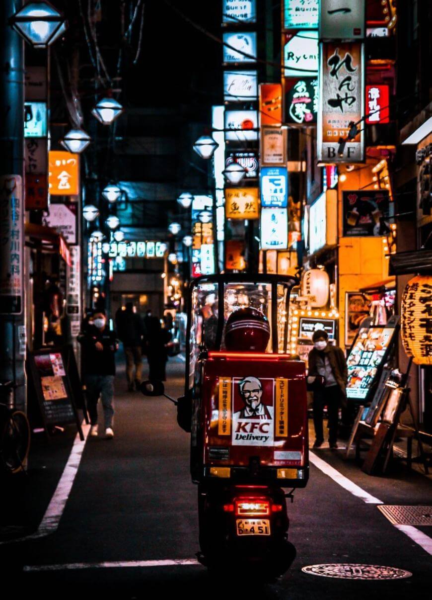 A KFC delivery scooter drives down a narrow, neon-lit street at night in a busy city, surrounded by colorful Japanese signs and people walking.