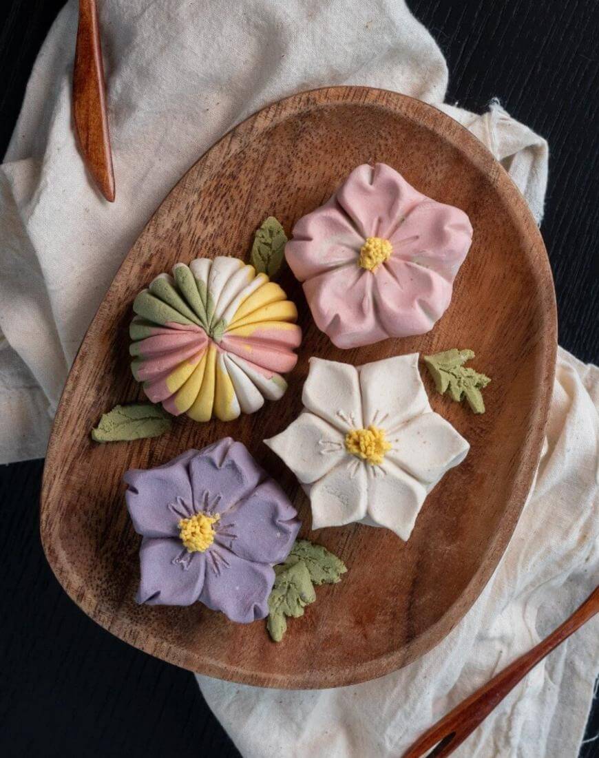 A wooden plate with four flower-shaped pastries in pastel colors—pink, white, purple, and yellow-green—decorated with leaf-shaped pieces, placed on a white cloth against a dark background.