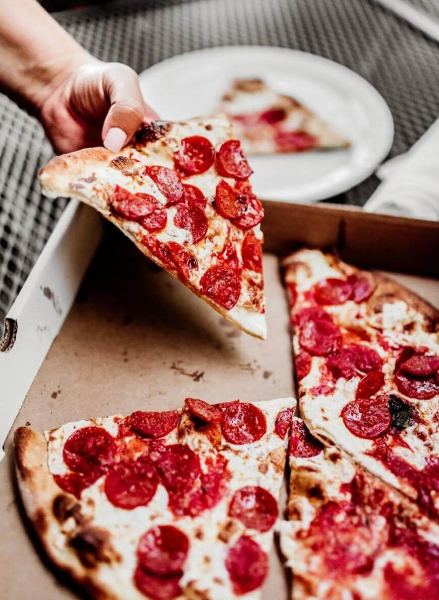 A hand lifts a slice of pepperoni pizza from a pizza box. Several remaining slices are in the box, and a single slice is on a white plate in the background.