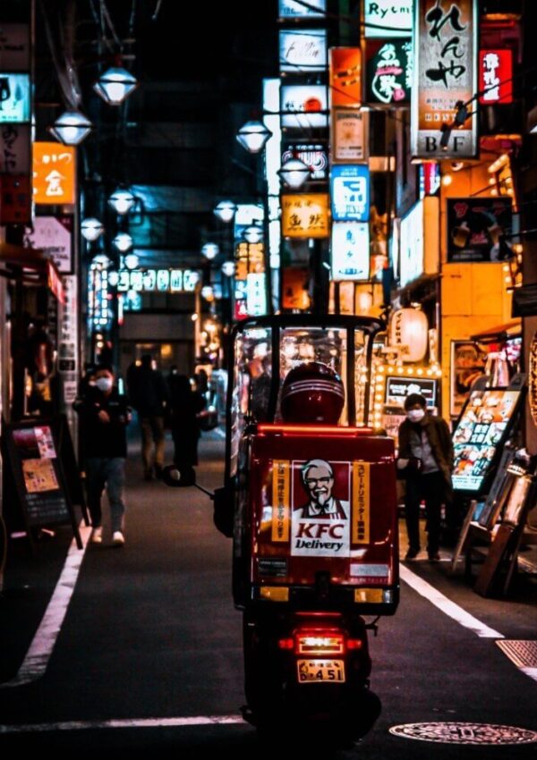 A KFC delivery scooter drives through a vibrant, neon-lit street lined with Japanese signs and restaurant fronts at night, with people walking and illuminated advertisements in the background.