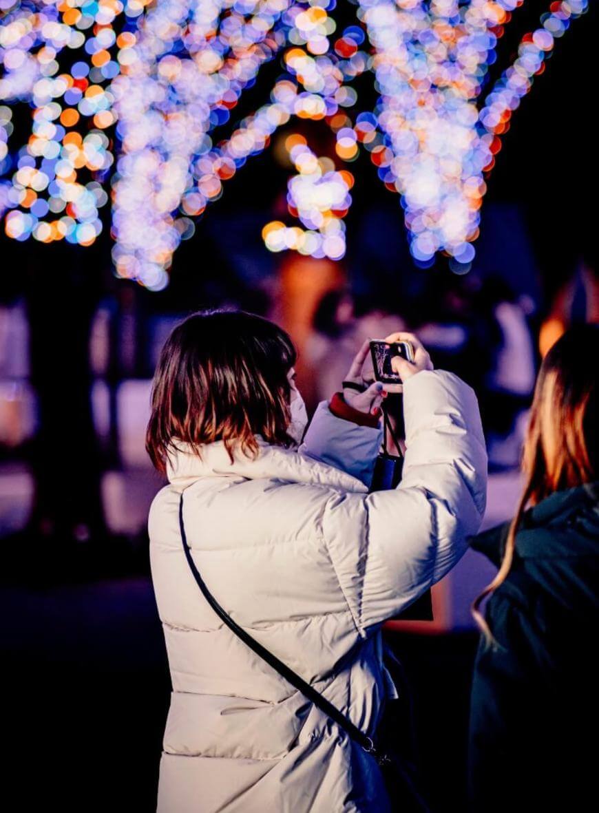 A person in a white puffy jacket takes a photo with a camera at night, standing next to another person. Colorful, blurred holiday lights decorate tree trunks in the background.