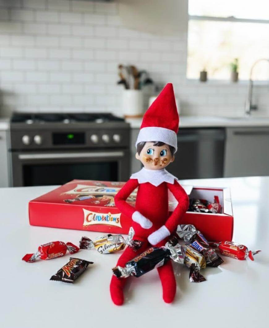 A toy elf in a red outfit sits on a kitchen counter with a red box of Celebrations chocolates behind it. Unwrapped chocolates are scattered around the elf. The kitchen has white tiles and stainless steel appliances.