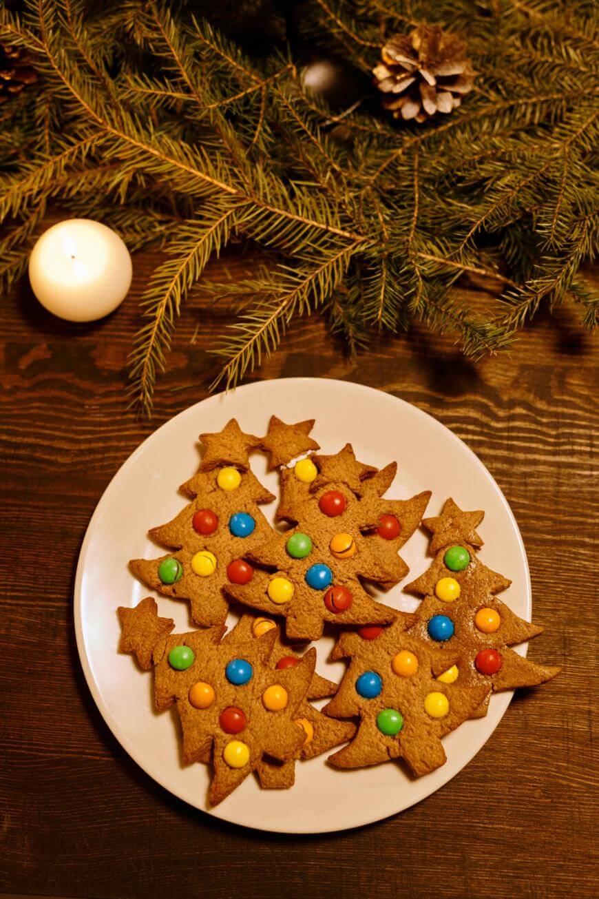 A white plate with Christmas tree-shaped cookies decorated with colorful candies sits on a wooden table. A lit candle, pinecones, and evergreen branches add a festive touch in the background.