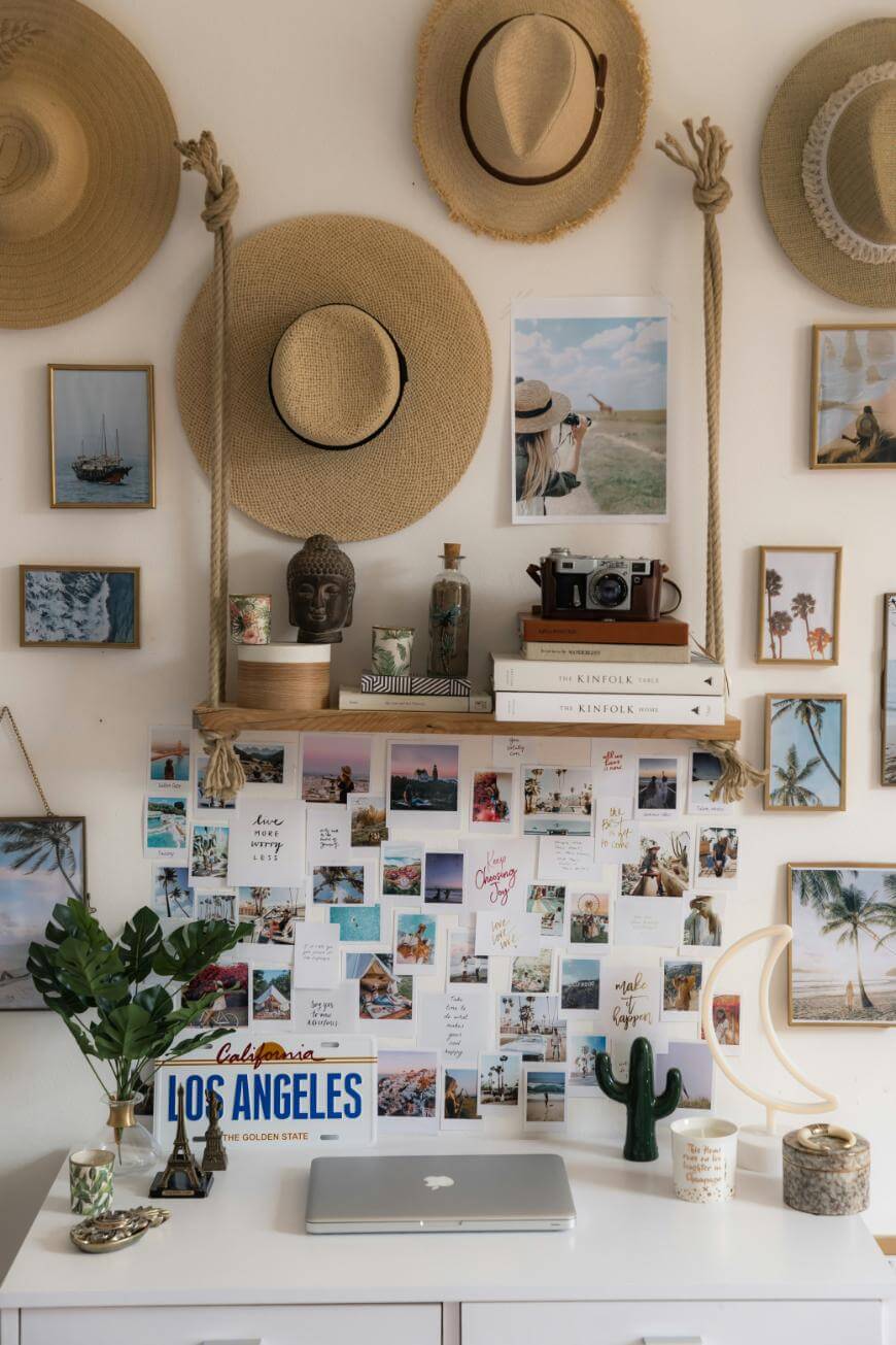 A decorated desk with a closed laptop, plants, candles, and a Los Angeles license plate. The wall above is covered with hats, photos, art, postcards, and books, creating a cozy, travel-inspired atmosphere.