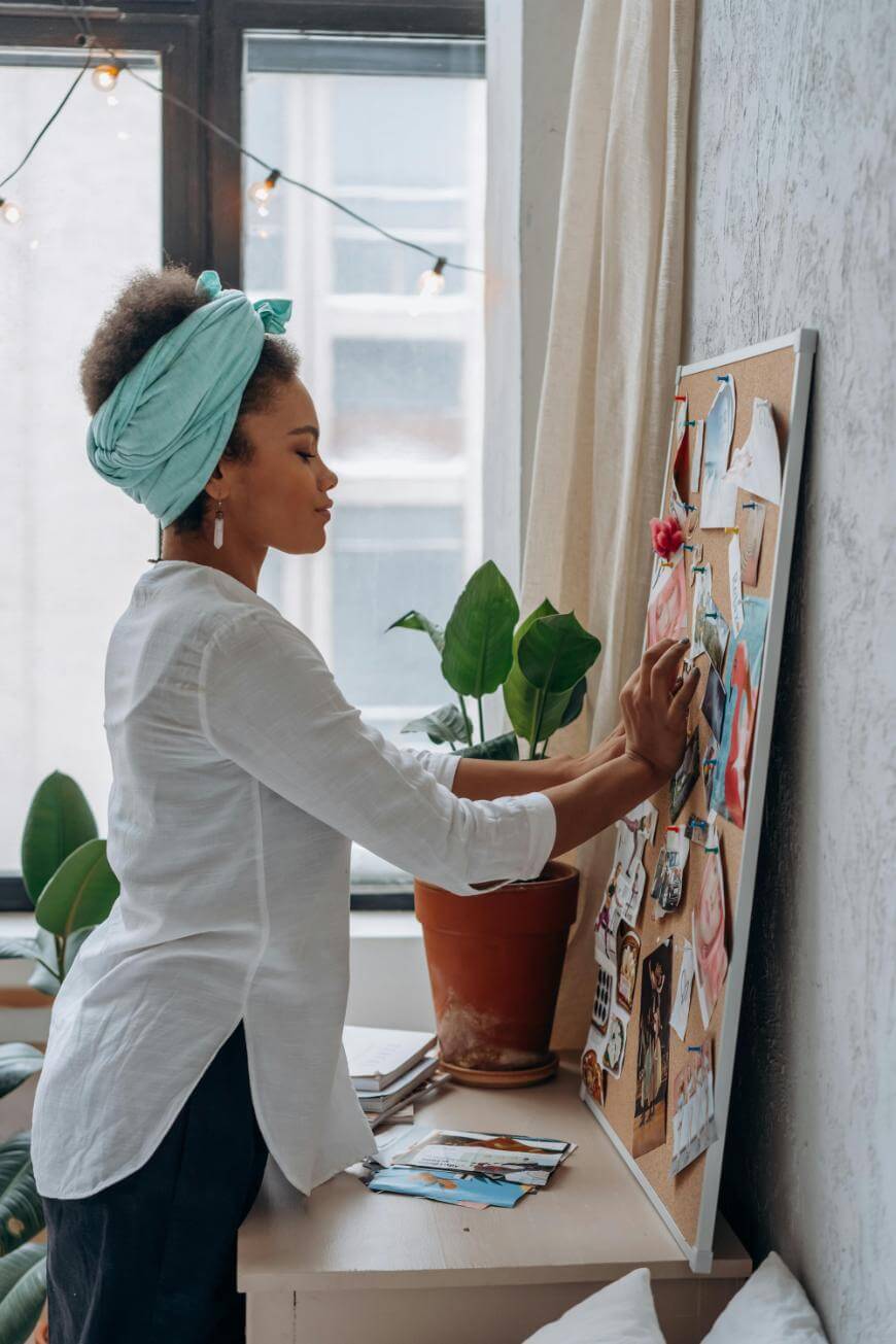 A woman wearing a teal headwrap and white shirt stands by a window, pinning photos onto a corkboard. A potted plant sits on the desk near her, and string lights hang in the background.
