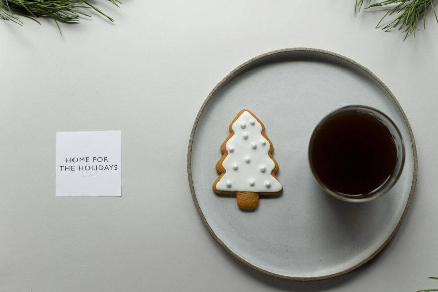 A plate with a tree-shaped iced cookie and a glass of dark drink sits next to a card that reads “Home for the Holidays”; sprigs of greenery are in the corners.