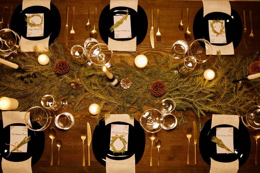 A festive table set for a meal, with black plates, cutlery, glasses, beige napkins, and menus. The centerpiece features pine branches, pinecones, and candles, creating a warm, rustic atmosphere.