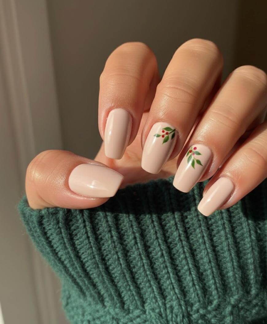 A hand with short, square-shaped nails painted in a soft nude shade. Two nails feature simple green leafy designs with small red dots. The person is wearing a green knit sweater.