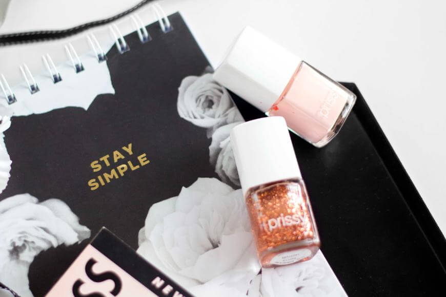 A flat lay image featuring a black notebook with STAY SIMPLE written on the cover, two nail polish bottles (one pale pink, one glittery gold), and a partially visible spiral notepad with floral designs.