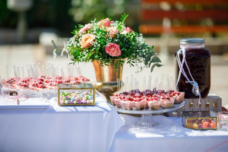 A decorated dessert table with assorted sweets in glass dishes, a large jar of dark beverage, and a gold vase filled with pink and peach flowers, all arranged on a white tablecloth outdoors.