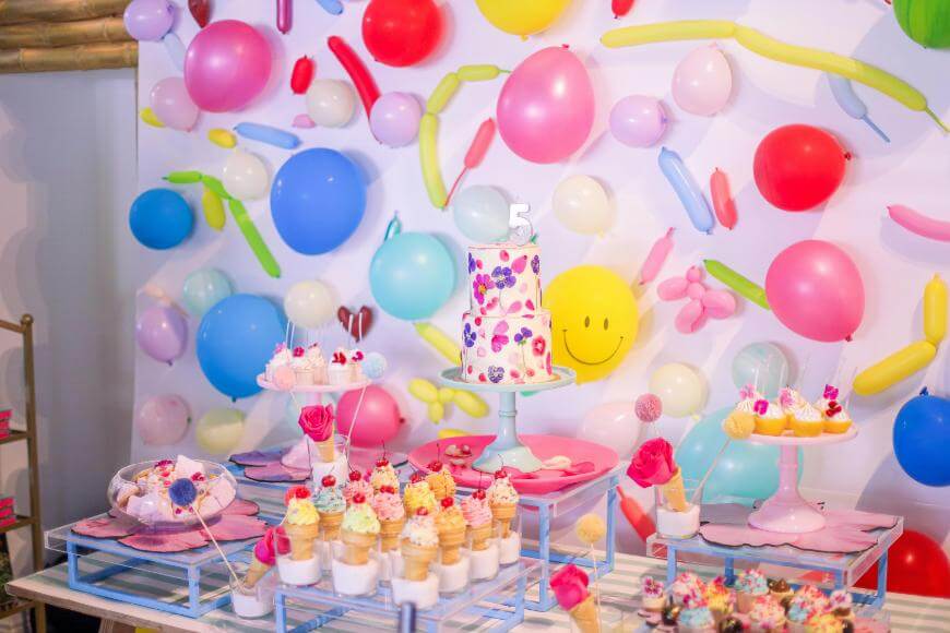 A colorful birthday party table with a decorated cake, cupcakes, and treats. The background is filled with bright balloons in various colors and shapes, including a yellow smiley face balloon.