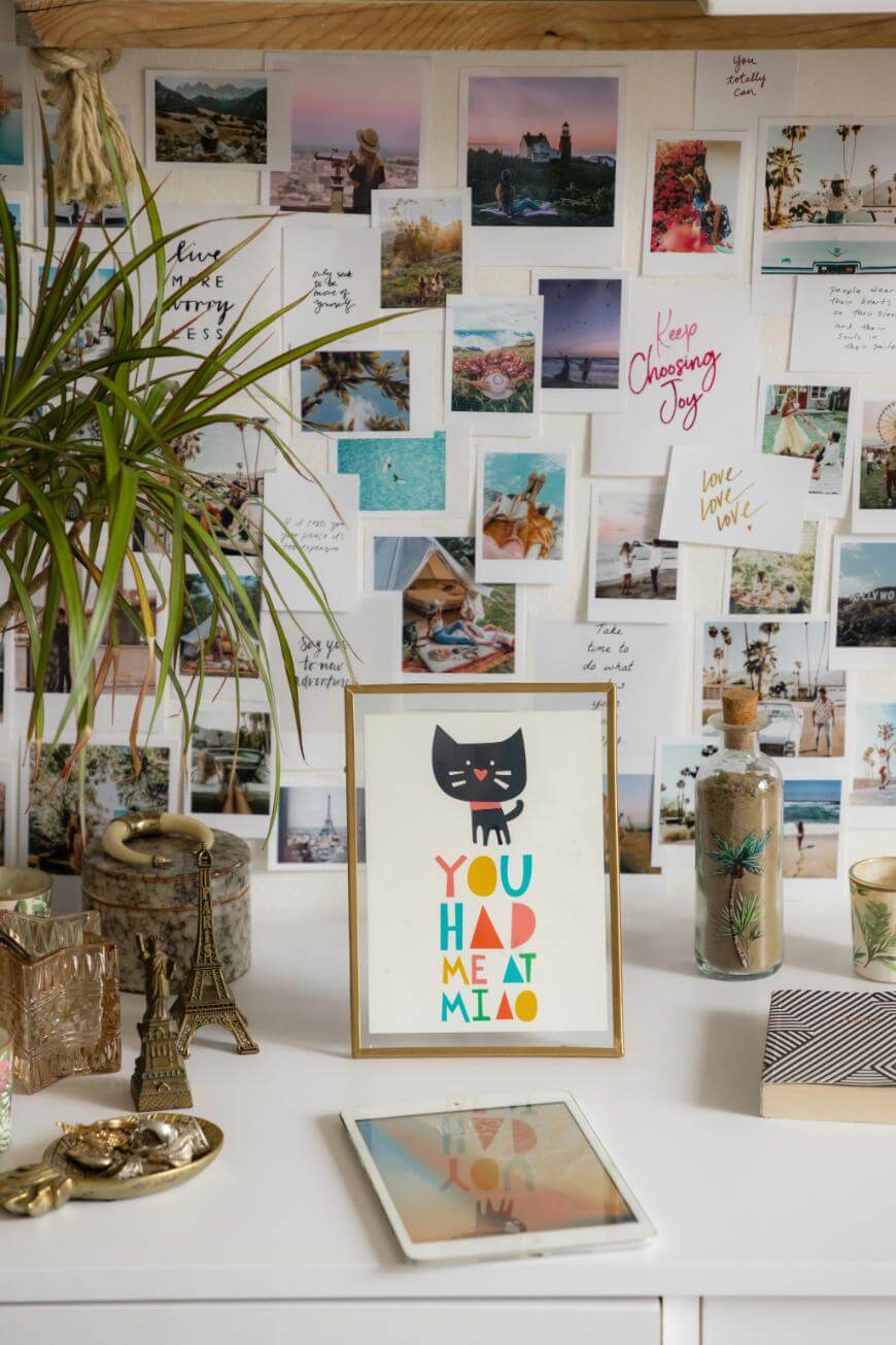 A desk with a framed print reading “You had me at meow” featuring a black cat, various decorative items, a potted plant, and an iPad. The wall behind is covered with travel photos and positive handwritten notes.