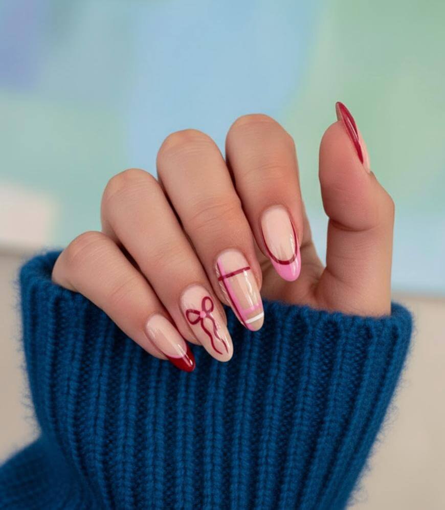 A hand with long, almond-shaped nails features a pink and red manicure with abstract and line designs, including a bow on the ring finger. The person wears a blue knit sweater.