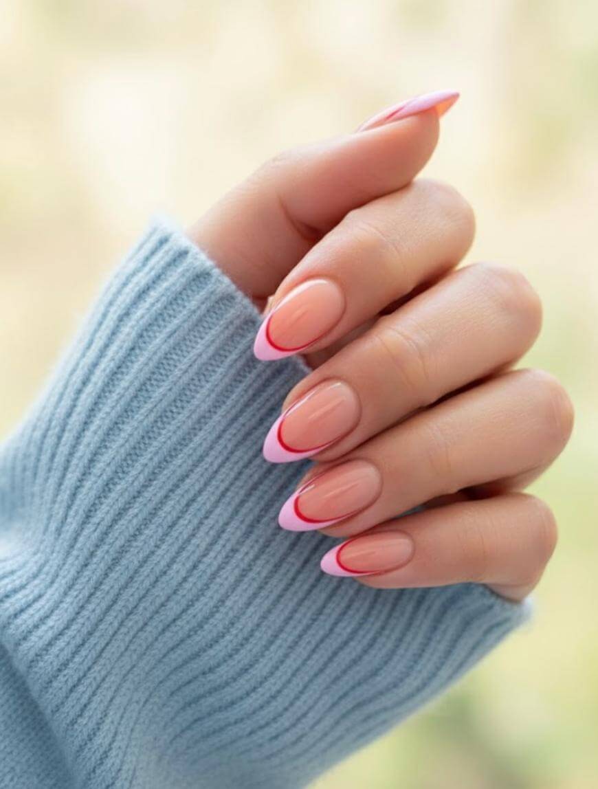 A hand with almond-shaped nails featuring a nude base and pink tips outlined in red, partially covered by the sleeve of a light blue knitted sweater.
