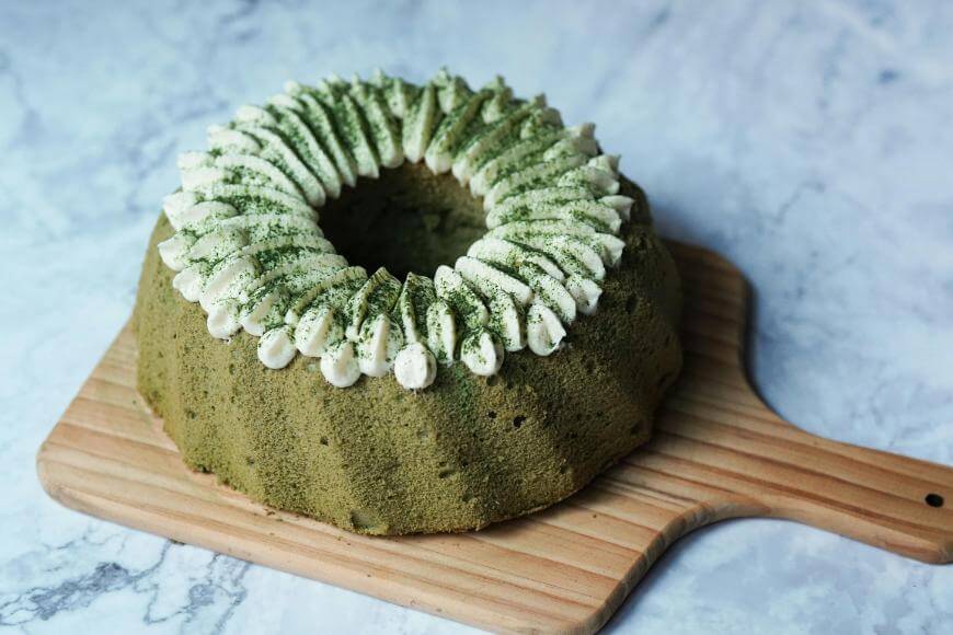A green matcha chiffon cake with white cream and powdered matcha on top, displayed on a wooden cutting board against a marble background.