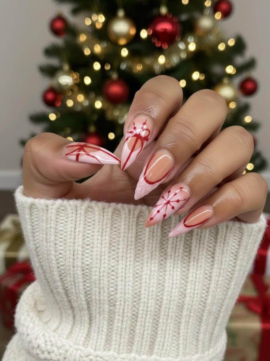 A hand with long, almond-shaped nails featuring festive red and pink snowflake nail art, held in front of a decorated Christmas tree with red and gold ornaments. The person is wearing a cream knit sweater.