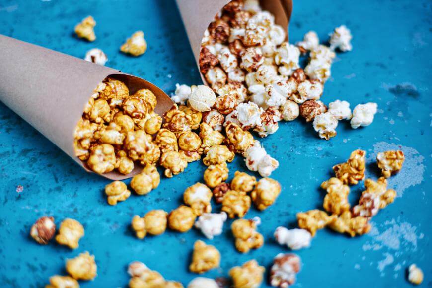 Two paper cones filled with popcorn, one with caramel popcorn and the other with regular popcorn, are lying on a blue surface with some popcorn scattered around them.