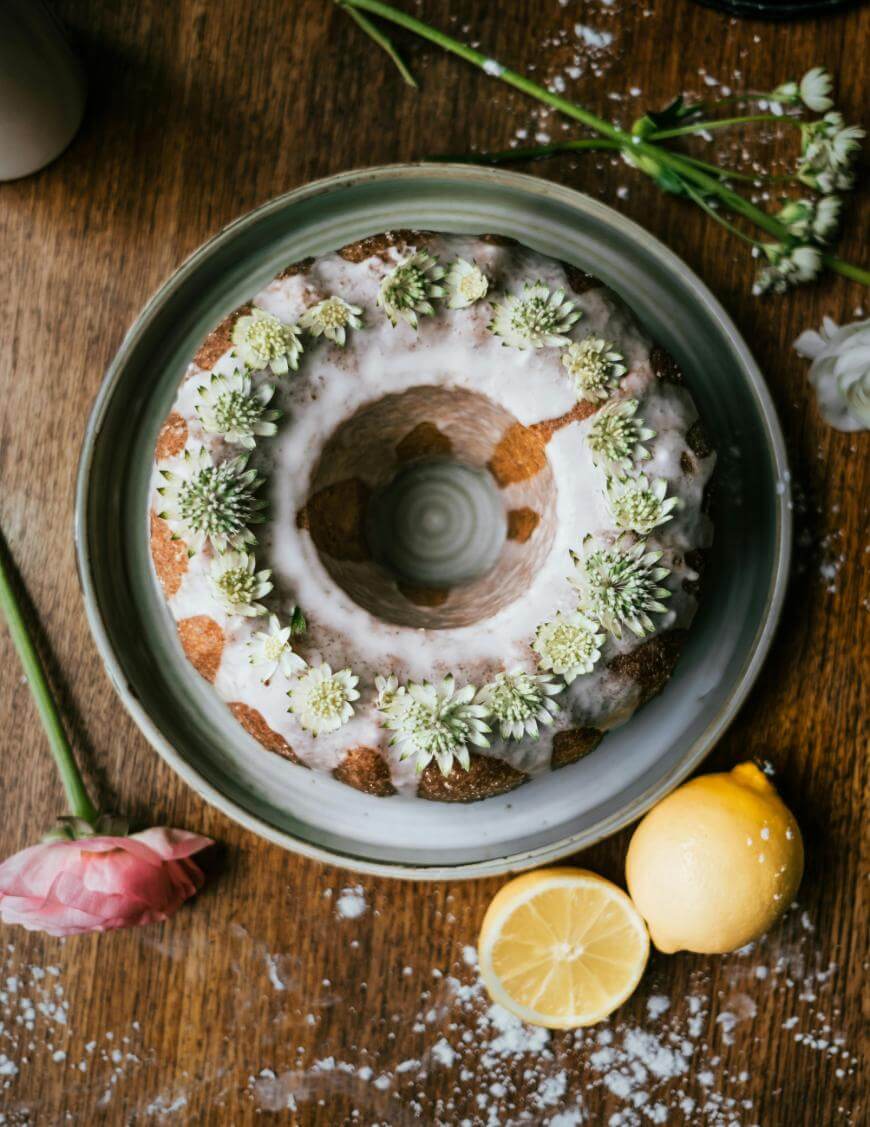 A bundt cake with white icing and small white flowers on top sits on a plate. A whole lemon, a lemon half, and scattered flowers rest on a wooden surface nearby.