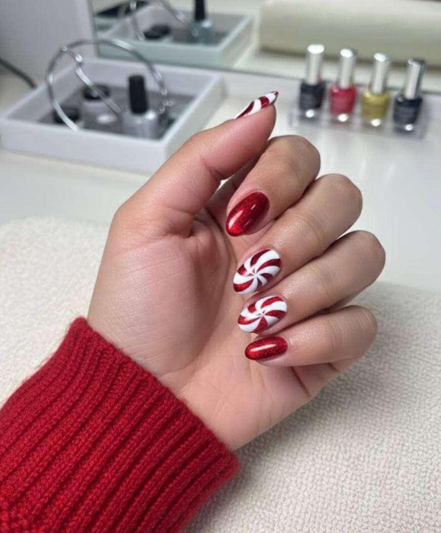 A hand with festive red and white nail art, featuring candy cane and peppermint swirl designs, rests on a towel. The person wears a red sweater, and bottles of nail polish are visible in the background.