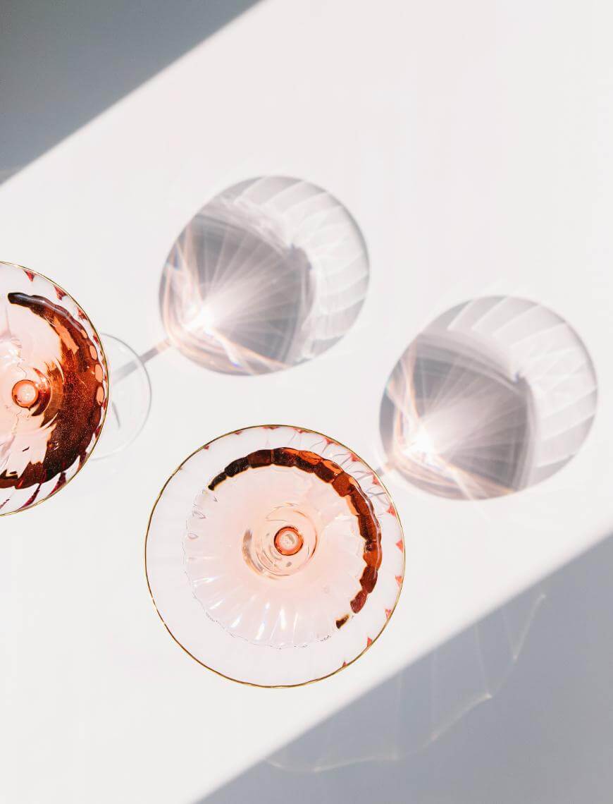 Two glasses of rosé wine viewed from above cast intricate shadows and reflections on a white surface, with light and geometric patterns created by the glass and liquid.