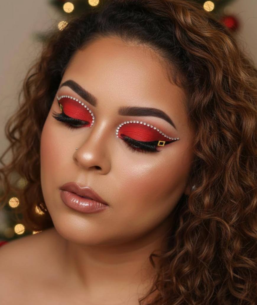 A woman with curly hair wears bold holiday eye makeup resembling Santa suits, with red lids, black liner, gold buckle designs, and white pearl accents. Her eyes are closed, and she has glossy nude lips.