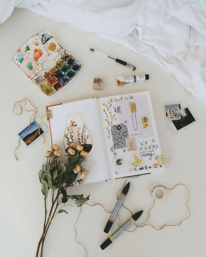 An open art journal with sketches, photos, and quotes is surrounded by dried flowers, watercolor paints, art supplies, twine, and loose photos on a white surface.