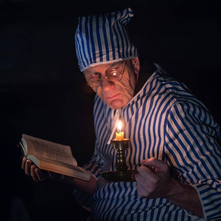 An older man wearing blue-and-white striped pajamas and a matching nightcap holds a candle and an open book. He looks serious and wears round glasses, sitting in a dimly lit, dark setting.