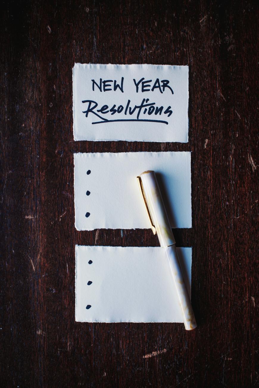 A piece of paper with “New Year Resolutions” written at the top, two blank sheets below with bullet points, and a pen resting on the bottom sheet, all placed on a dark wooden surface.