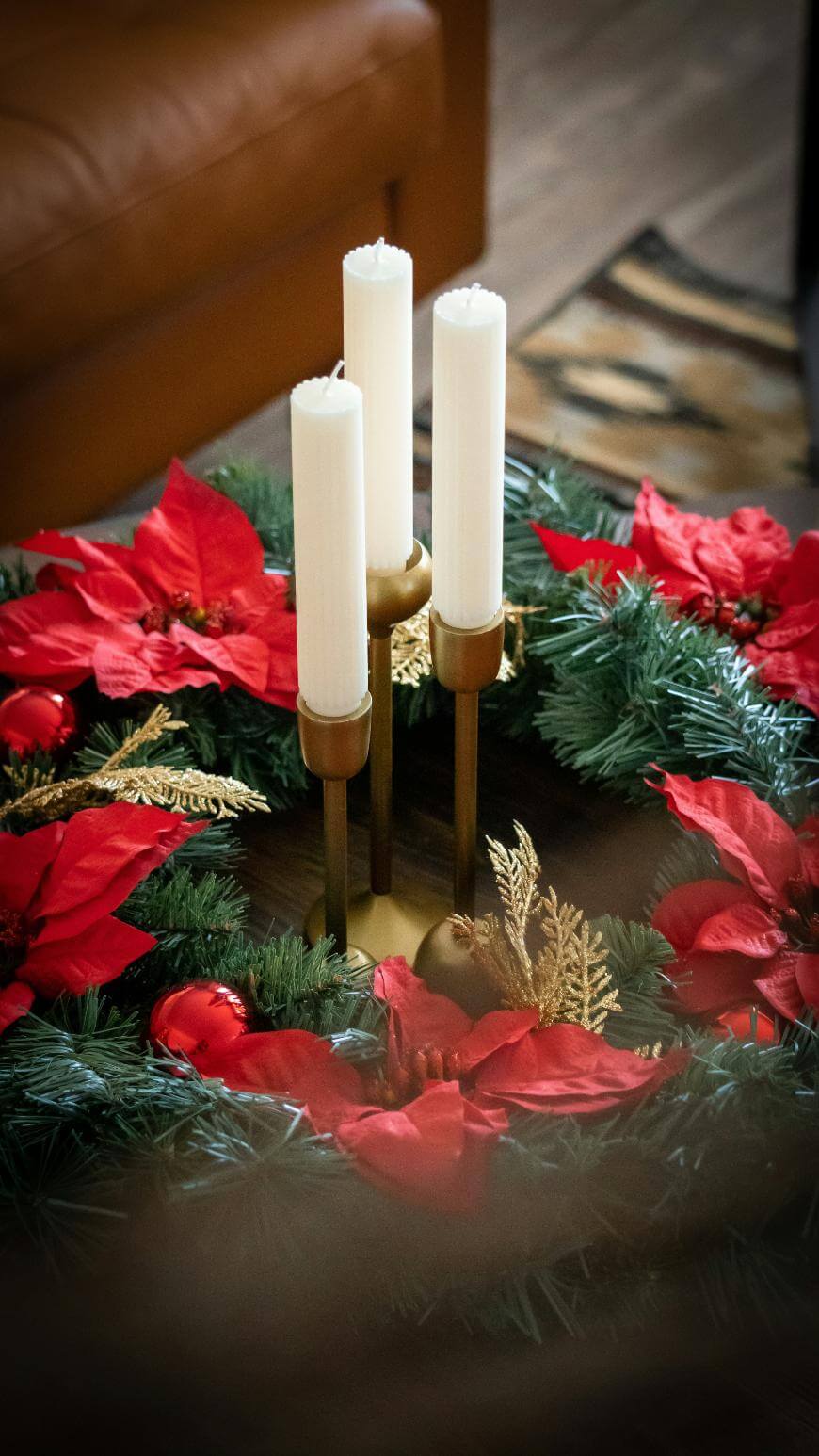 A festive wreath with red poinsettias, green pine branches, and gold accents surrounds three white candles in gold holders, set on a table with a blurred background.