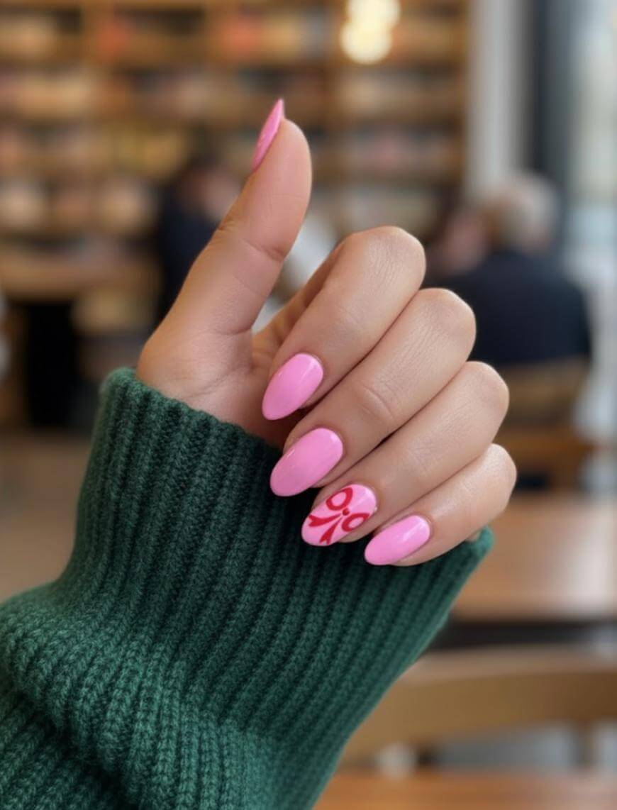 A hand with pink manicured nails, one featuring a red design, is shown against a blurred background. The person is wearing a dark green, ribbed sweater.