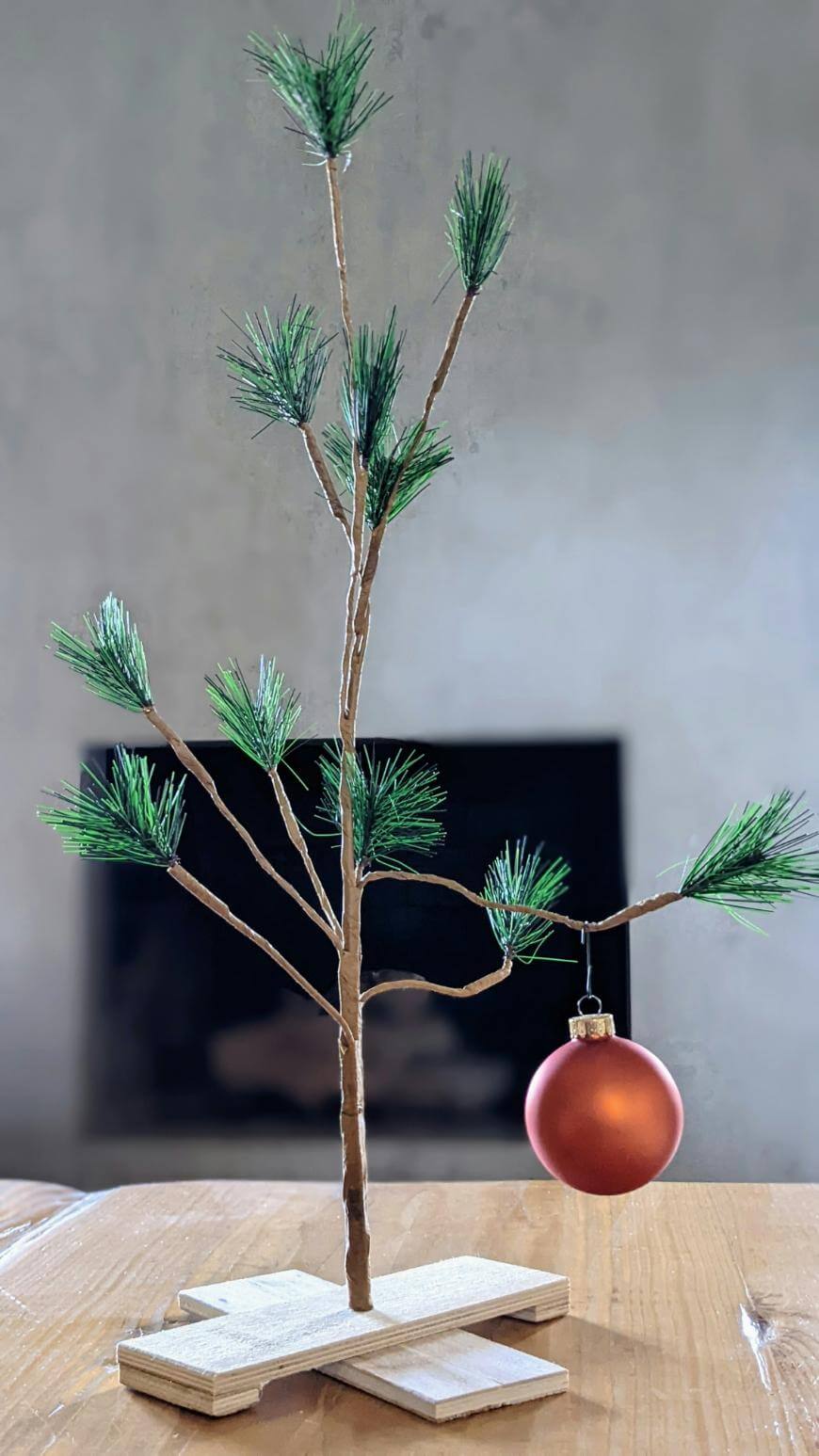 A small, sparse artificial Christmas tree with a few green branches stands on a wooden base. A single red ornament hangs from one of the lower branches. The background is softly blurred.