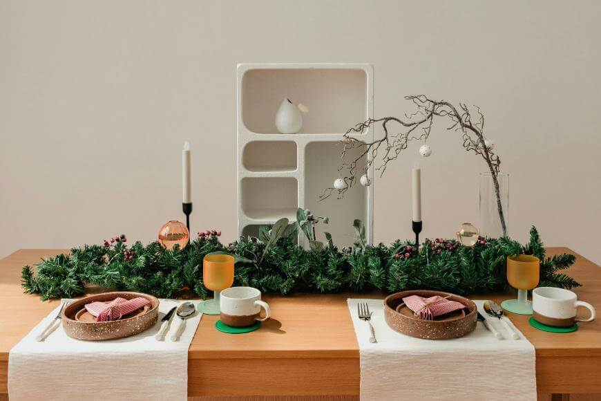 A festive table is set for two with white plates, brown bowls, pink napkins, gold-rimmed glasses, and white mugs. A green garland with candles and ornaments decorates the center. A modern white shelf is in the background.