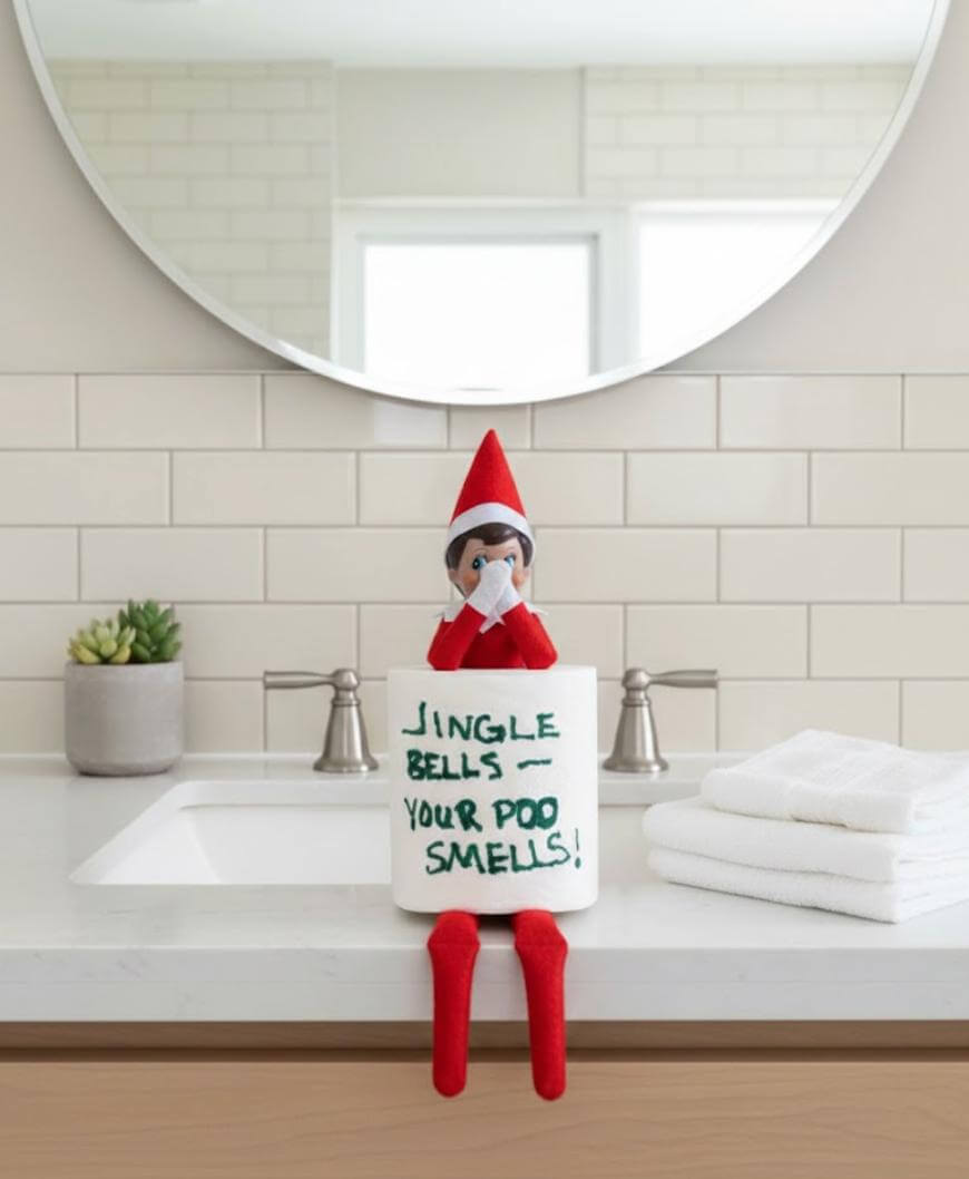 An Elf on the Shelf doll sits on a bathroom counter holding a toilet paper roll that says, “Jingle Bells — Your Poo Smells!” The elf faces forward, with a sink, mirror, and folded towels in the background.