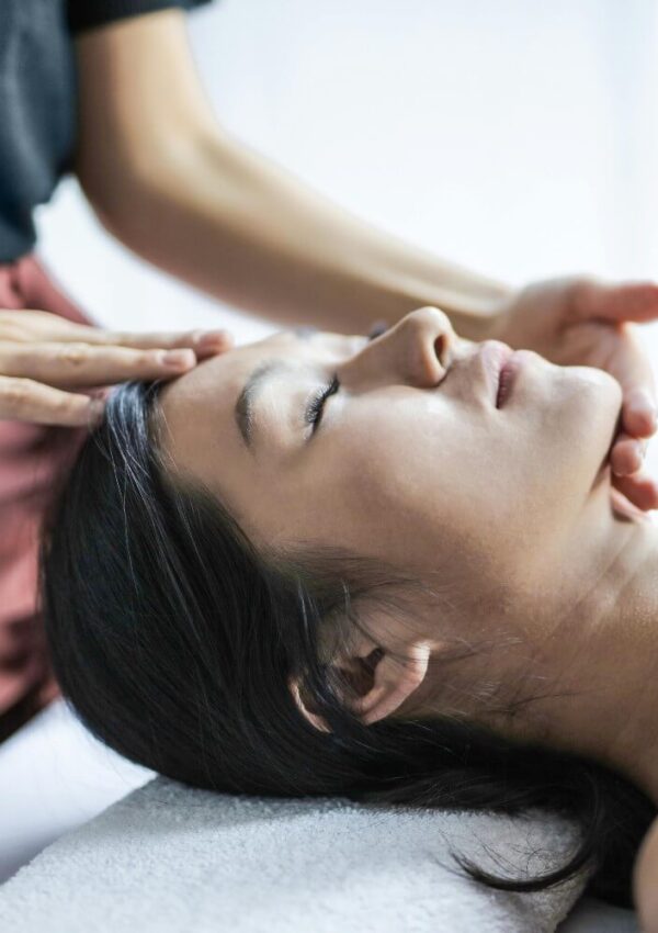 A person with long dark hair lies with eyes closed on a white towel while another gently massages their forehead and jaw, suggesting a relaxing facial massage or skin rejuvenation at Treat Medspa NYC.