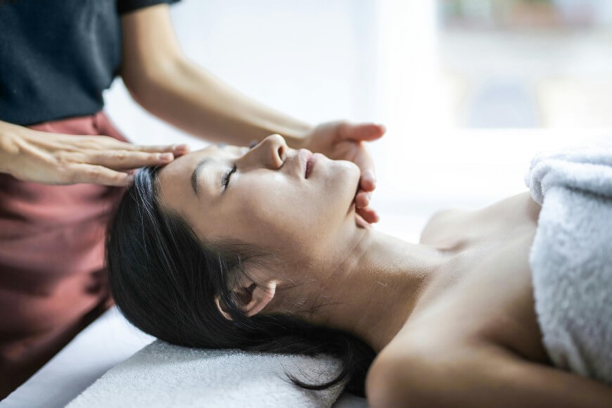 A person lies on a massage table with eyes closed, covered by a white towel, while another gently massages their forehead and jaw in a calm, bright Medspa NYC setting focused on relaxation and skin rejuvenation.