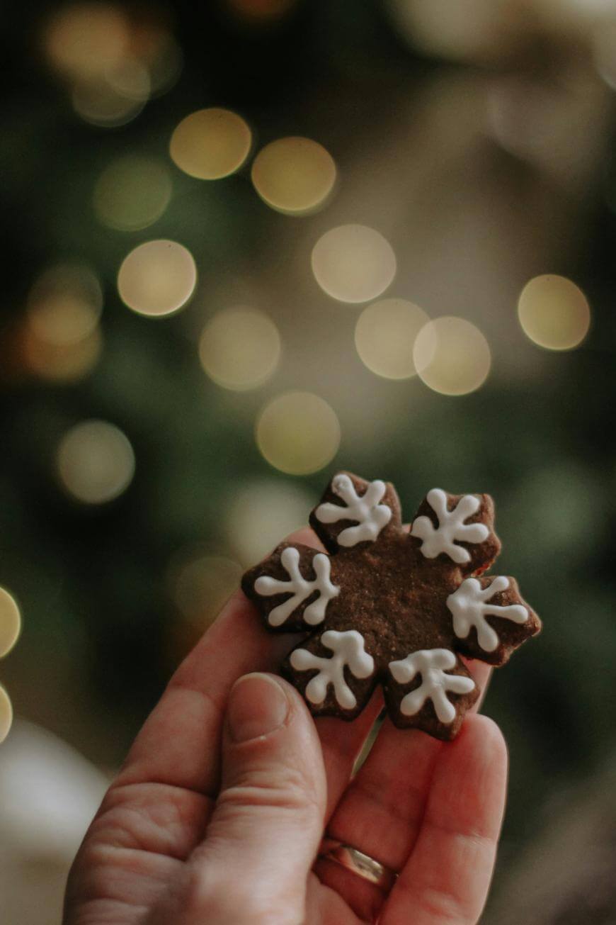 A hand holding a brown, snowflake-shaped cookie decorated with white icing, with blurred warm lights and greenery in the background.