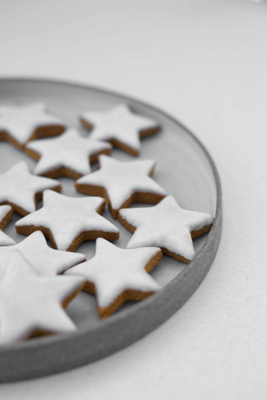 A grey plate with star-shaped cookies topped with white icing, arranged on a light surface. Some cookies are partially off the plate, and the image has a minimalistic, clean background.