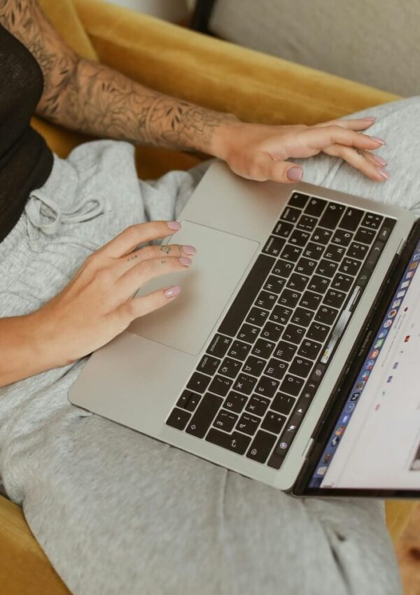 A person with a tattooed arm and pink nails sits on a chair in gray sweatpants, using a laptop on their lap.