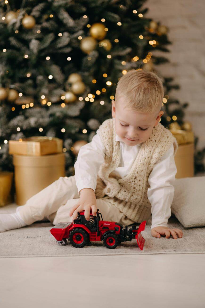 A young boy in light-colored clothes sits on the floor by a Christmas tree, playing with a red toy tractor. Golden ornaments and gift boxes are visible under the decorated tree in the background.